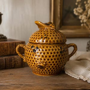 May include: A round, golden-brown ceramic honey pot with a textured honeycomb pattern and two side handles. The lid features a sculpted bee handle, and small dark bee figures are embossed on the surface of the pot, creating a detailed, tactile design.