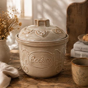 Peut inclure: Un pot à biscuits en céramique beige avec un couvercle assorti, orné de motifs en relief en forme de cœur et de bretzel sur le dessus et du mot Cookies en écriture cursive sur le devant. Le pot a une finition brillante et une forme arrondie.