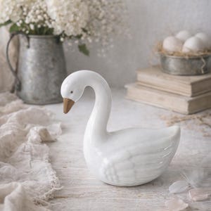 Può includere: Una statuetta di cigno in ceramica bianca con collo curvo e becco marrone. Il cigno è su una superficie di legno bianco, con un vaso di fiori bianchi e un piccolo contenitore di metallo con uova sullo sfondo. Arredamento per la casa.