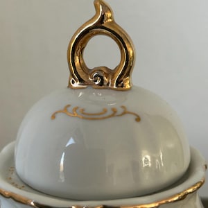 May include: Close-up of a white porcelain lid with gold trim and a gold handle. The lid has a rounded shape with a gold decorative design. The handle is a loop shape with a decorative design.