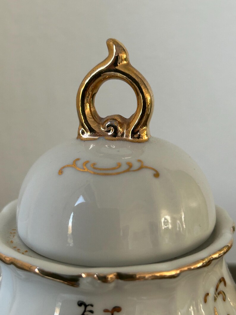 May include: Close-up of a white porcelain lid with gold trim and a gold handle. The lid has a rounded shape with a gold decorative design. The handle is a loop shape with a decorative design.