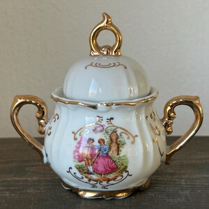 May include: White porcelain sugar bowl with a lid and gold accents. The bowl features a painted scene of two figures in a garden. The handles and lid finial are gold-colored. A decorative piece for serving sugar.
