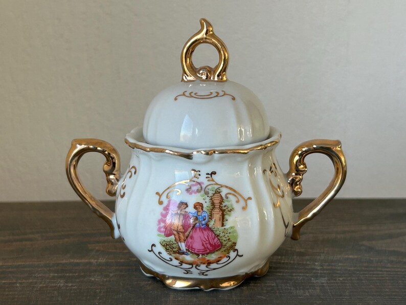 May include: White porcelain sugar bowl with a lid and gold accents. The bowl features a painted scene of two figures in a garden. The handles and lid finial are gold-colored. A decorative piece for serving sugar.