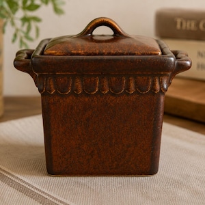 Pode incluir: Um recipiente quadrado de cerâmica castanho-escuro com tampa a condizer e duas pegas laterais. O recipiente apresenta um detalhe decorativo de rebordo recortado na parte superior e um acabamento vidrado com variações de cor.