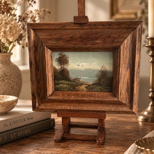 Op de afbeelding: Een klein landschapsschilderij in een dikke, donkerbruine houten lijst, tentoongesteld op een bijpassende houten ezel. Het kunstwerk toont een landelijk tafereel met een zandpad, bomen en twee vogels in een bleke lucht, in gedempte aardetinten.