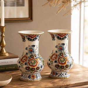 Op de afbeelding: Twee witte keramische vazen met een bloemenontwerp in oranje, blauw en groen. De vazen hebben een uitlopende bovenkant en een bolvormig lichaam, met een gedetailleerd patroon rond de basis en de bovenkant. Geplaatst op een houten oppervlak.