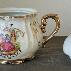 May include: Cream-colored porcelain sugar bowl with gold trim and handles, featuring a romantic scene with a couple in period clothing. The lid is also cream-colored with gold accents.
