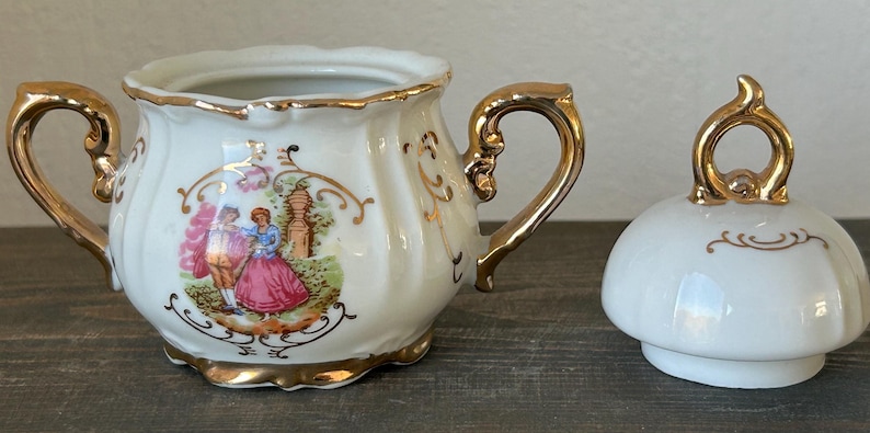 May include: Cream-colored porcelain sugar bowl with gold trim and handles, featuring a romantic scene with a couple in period clothing. The lid is also cream-colored with gold accents.
