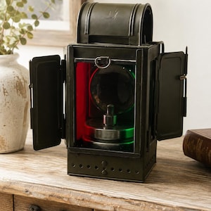 Può includere: Una lanterna in metallo verde oliva scuro con telaio rettangolare e maniglia superiore arrotondata. La lanterna presenta due porte laterali aperte, una lente in vetro circolare centrale e pannelli in vetro rosso e verde attorno a una base.