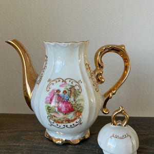 May include: White porcelain teapot with gold trim, handle, and spout. The teapot features a painted scene of figures in a garden and a matching lid. The teapot is on a dark wooden surface.