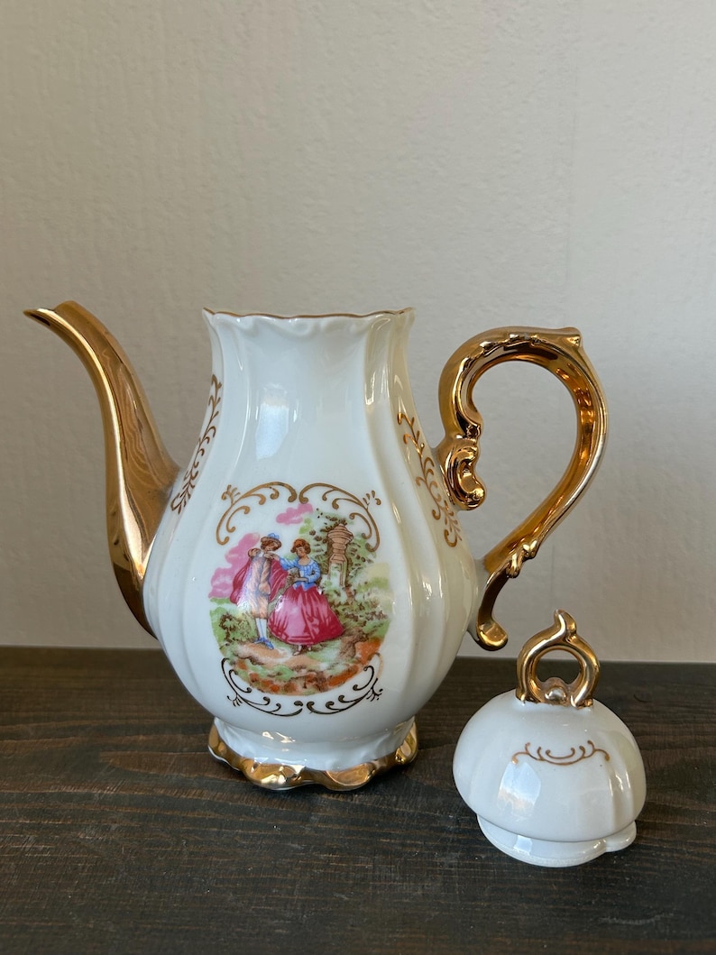 May include: White porcelain teapot with gold trim, handle, and spout. The teapot features a painted scene of figures in a garden and a matching lid. The teapot is on a dark wooden surface.