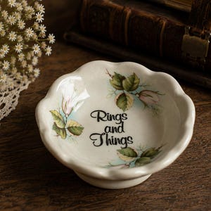 May include: A small, round, white ceramic trinket dish with a scalloped edge. The interior features a floral design with green leaves and pink buds, surrounding the black text "Rings and Things" printed in the center of the bowl.