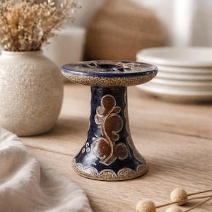Puede incluir: Un portavelas de cerámica con base azul oscuro y diseño floral marrón. La parte superior es azul oscuro con una hendidura circular. El portavelas está sobre una superficie de madera.