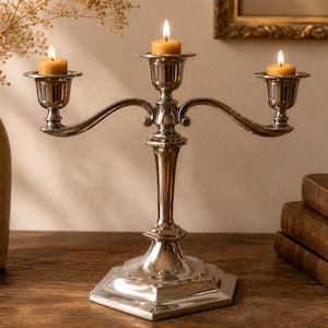 May include: A silver-toned metal candelabra featuring three arms with flared candle cups, each holding a small, lit, cylindrical yellow candle. The candelabra has a central stem and a hexagonal base, providing a stable foundation for the structure.