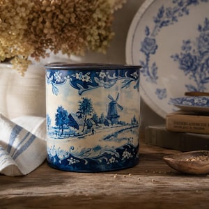 Op de afbeelding: Een cilindrische metalen bus met een blauw-wit transferware-patroon van een landelijk landschap met een windmolen, huizen en bomen, omlijst door decoratieve bloemmotieven op een crèmekleurige achtergrond met een donkerblauwe rand.