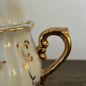 May include: Cream-colored porcelain sugar bowl with a gold-colored handle and rim. The handle is ornate, and the bowl features gold decorative accents. The lid is also cream-colored with a gold rim. A classic piece for serving sugar.