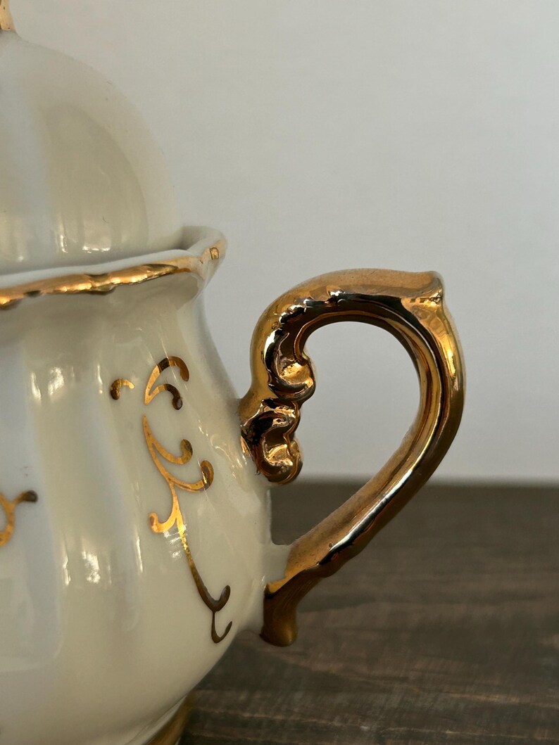 May include: Cream-colored porcelain sugar bowl with a gold-colored handle and rim. The handle is ornate, and the bowl features gold decorative accents. The lid is also cream-colored with a gold rim. A classic piece for serving sugar.