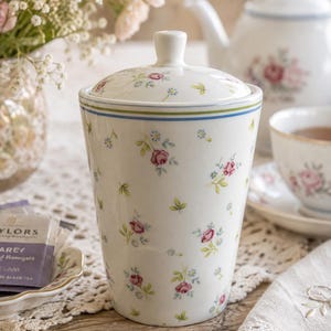 May include: A white ceramic canister with a lid, featuring a delicate floral pattern of small pink roses and tiny blue flowers. The canister has a tapered cylindrical shape with thin blue and green horizontal stripes near the top rim.