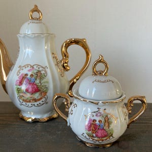 May include: A white porcelain tea set with gold accents. The set includes a teapot and a sugar bowl, both featuring a romantic scene in a decorative frame. The teapot has a curved handle and a lid with a gold finial.