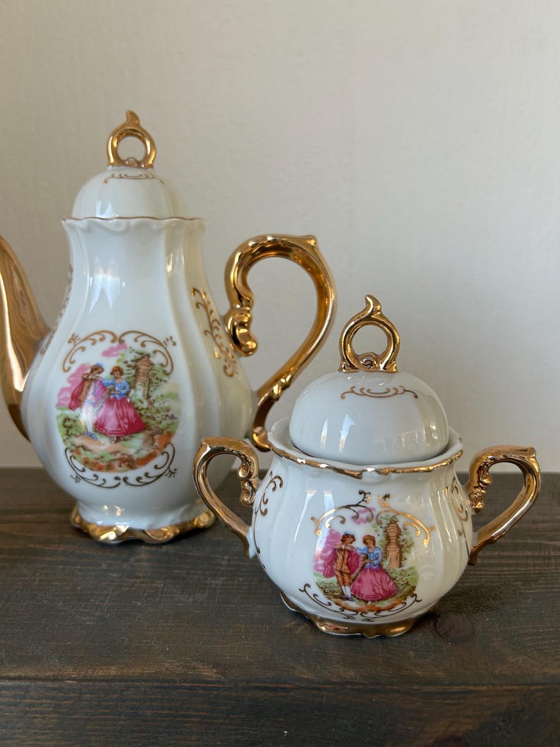 May include: A white porcelain tea set with gold accents. The set includes a teapot and a sugar bowl, both featuring a romantic scene in a decorative frame. The teapot has a curved handle and a lid with a gold finial.