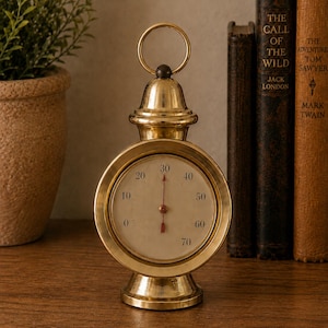May include: A polished brass thermometer with a circular face, a cream-colored dial, and black numerical markings ranging from 0 to 70. The device features a central red indicator needle, a decorative top ring for hanging, and a sturdy circular base.