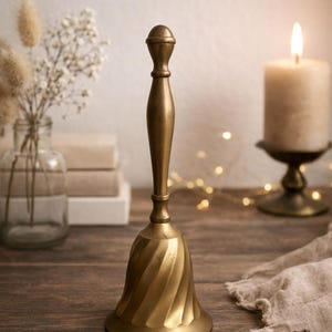 Op de afbeelding: Een messing bel met een spiraalvormig ontwerp op de bel en een lange handgreep. De bel staat op een houten oppervlak, met een brandende kaars en een vaas met bloemen op de achtergrond. Decoratief item.