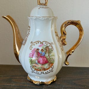 May include: White porcelain teapot with gold trim and handle. The teapot features a painted scene of a couple in a garden, framed by gold filigree. The lid has a gold loop handle. The spout and base are also trimmed in gold.