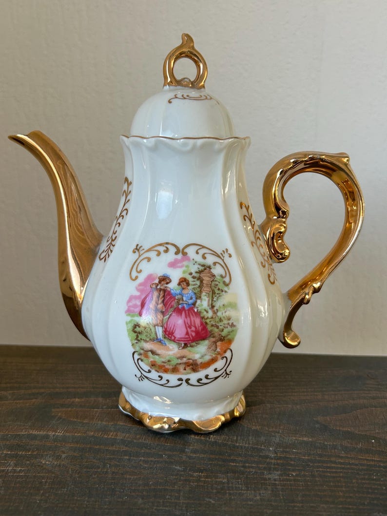 May include: White porcelain teapot with gold trim and handle. The teapot features a painted scene of a couple in a garden, framed by gold filigree. The lid has a gold loop handle. The spout and base are also trimmed in gold.