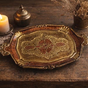 May include: An oval-shaped decorative tray featuring a distressed gold and deep red finish with an intricate embossed filigree pattern in the center. The tray has two ornate handles on the sides and a raised, scalloped edge with a textured metallic surface.