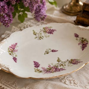 Könnte beinhalten: Eine ovale Servierplatte aus Porzellan mit weißem Grund, verziert mit einem zarten Blumenmuster aus violetten und rosafarbenen Fliederclustern. Die Platte hat einen gewellten Rand mit strukturierten, goldfarbenen Details an Griffen und Rand.