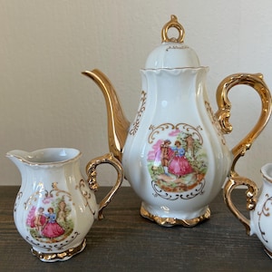May include: A white porcelain tea set with gold trim. The set includes a teapot, creamer, and sugar bowl, each featuring a romantic scene with figures in period clothing. The teapot has a curved spout and a decorative handle. The set is on a dark wooden surface.