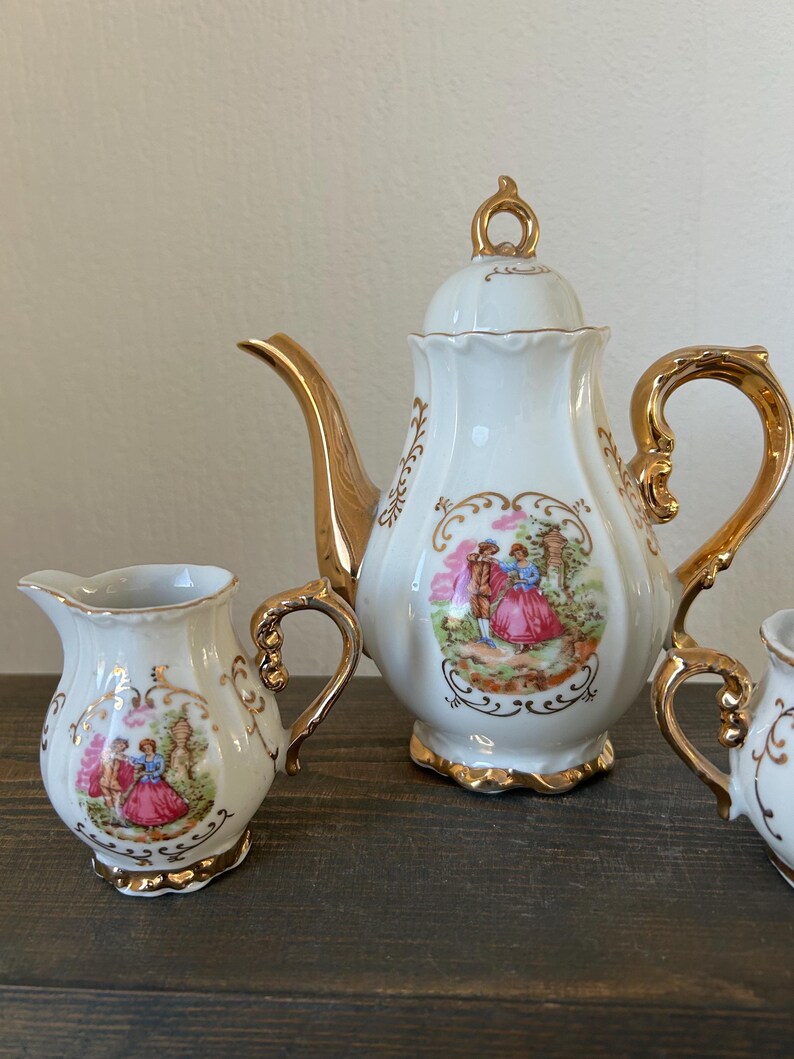 May include: A white porcelain tea set with gold trim. The set includes a teapot, creamer, and sugar bowl, each featuring a romantic scene with figures in period clothing. The teapot has a curved spout and a decorative handle. The set is on a dark wooden surface.