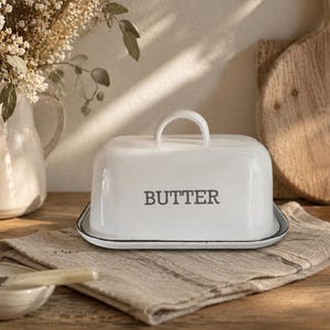 以下が含まれることがあります： 蓋と取っ手が付いた白いバター皿で、前面に「BUTTER」の文字がプリントされています。 皿はベージュのリネンのナプキン上にあり、背景には小さなボウルと木製のまな板があります。 ドライフラワーの花瓶も見える。