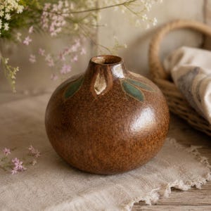 Pode incluir: Um vaso de cerâmica castanho de forma redonda com um acabamento texturizado e salpicado, e um pescoço estreito. A parte superior apresenta motivos de folhas verdes pintadas à volta da abertura, com um verniz brilhante sobre a base.