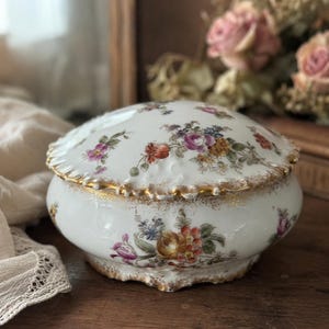 Op de afbeelding: Een witte porseleinen kom met deksel, voorzien van een handgeschilderd bloemmotief met roze, oranje en blauwe bloemen. De kom heeft een geschulpte rand met gouden accenten en een bijpassend deksel met vergelijkbare florale decoraties.