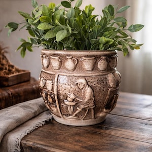 Może przedstawiać: Brązowa ceramiczna donica z misternym reliefem przedstawiającym postacie i ceramikę. Donica jest wypełniona zielonymi roślinami liściastymi. Projekt obejmuje małe dzbanki wokół górnej krawędzi.