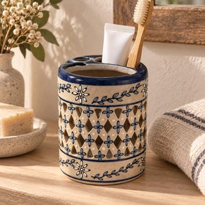 Puede incluir: Un soporte cilíndrico de cerámica para cepillos de dientes con base color crema y un patrón floral azul pintado a mano. El cuerpo presenta un diseño de celosía calada en forma de diamante. Contiene un tubo de pasta dental blanco y un cepillo de madera.