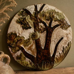 Pode incluir: Uma placa de parede circular de cerâmica com uma representação em relevo de uma árvore com um tronco castanho escuro e folhagem verde texturizada. O fundo é de cor creme esbranquiçada marmoreada com uma textura de superfície irregular.