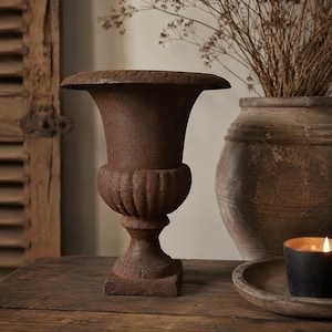 May include: A dark brown, weathered cast iron urn with a wide, flared rim, a fluted midsection, and a square base. The surface has a textured, rusted appearance with visible signs of age and oxidation, standing upright on a flat surface.
