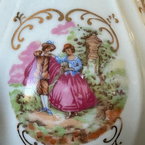 May include: A white porcelain decorative piece with a painted scene of a couple in a garden. The man wears a brown coat and the woman a pink dress. Gold filigree frames the scene.