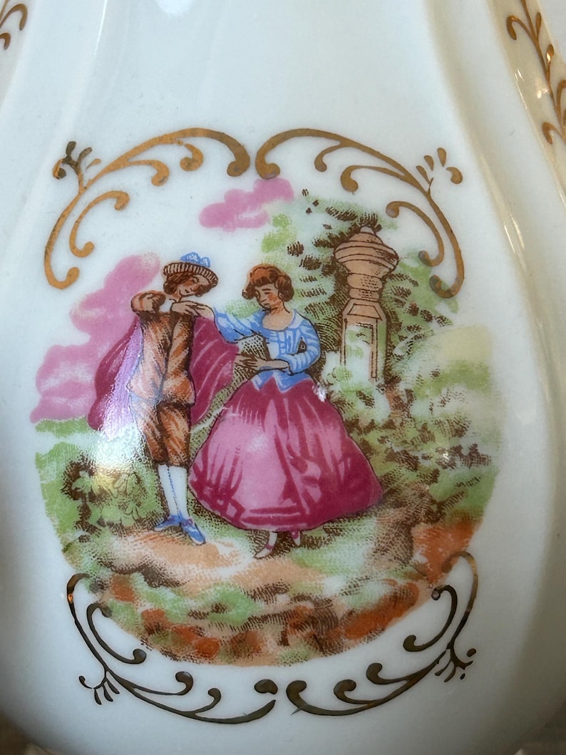May include: A white porcelain decorative piece with a painted scene of a couple in a garden. The man wears a brown coat and the woman a pink dress. Gold filigree frames the scene.