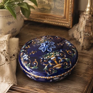 Könnte beinhalten: Eine runde, tiefblaue Keramikdose mit Deckel, verziert mit detaillierten, mehrfarbigen Blumen- und Rankenmustern. Der Deckel ist mit einem skulpturalen, dreidimensionalen blauen Blumenknauf versehen, und der Boden hat eine gelb-weiße Bordüre.