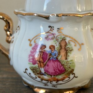 May include: A white porcelain tea set with gold trim and a lid. The tea set features a painted scene of a couple in a garden. The handle and edges are accented with gold. The tea set is on a wooden surface.