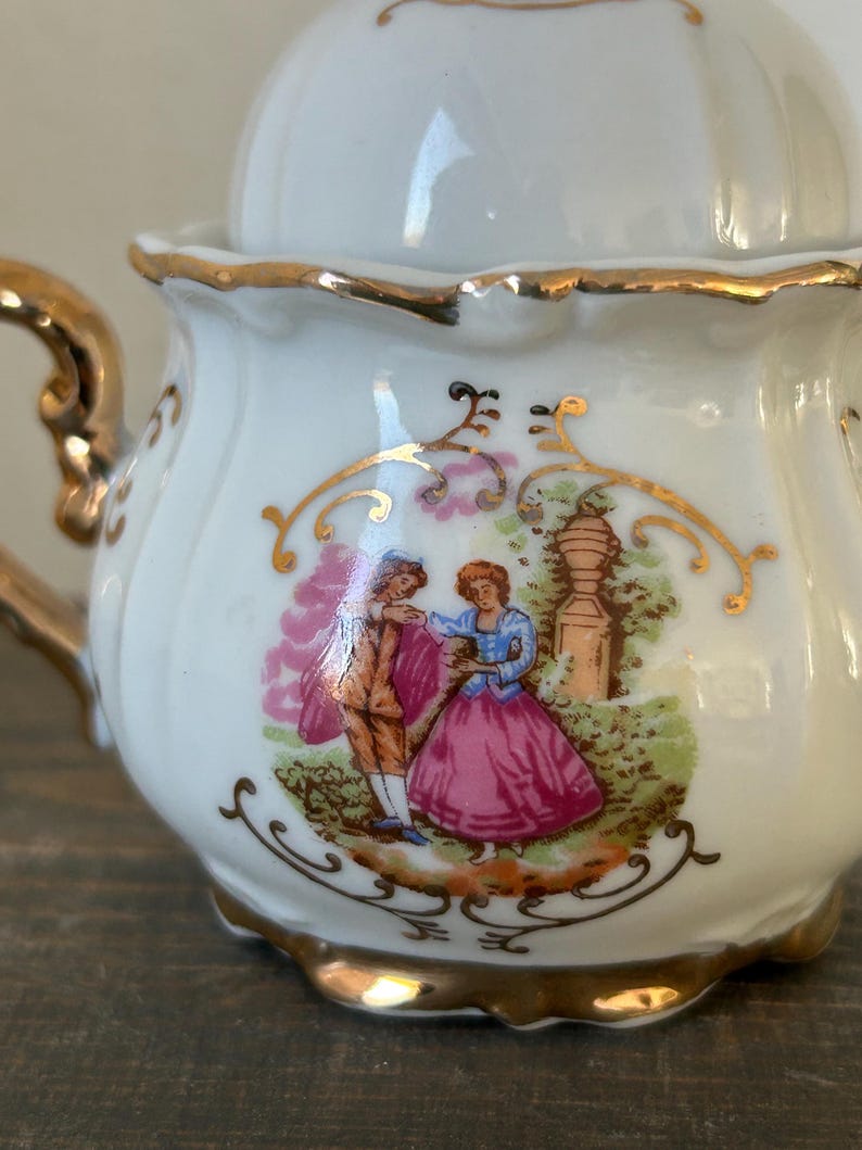 May include: A white porcelain tea set with gold trim and a lid. The tea set features a painted scene of a couple in a garden. The handle and edges are accented with gold. The tea set is on a wooden surface.