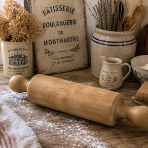 May include: A wooden rolling pin with rounded handles positioned horizontally on a surface dusted with white flour. The rolling pin features a smooth, light brown grain and a cylindrical body, set against a kitchen scene with various ceramic containers.