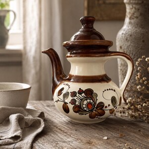 Può includere: Una teiera in ceramica con base color crema e dettagli smaltati marrone scuro sul coperchio, sul manico e sul beccuccio. Il corpo presenta un motivo floreale dipinto a mano nei toni del marrone, arancione e verde, con una forma arrotondata.