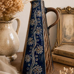 May include: A tall, conical ceramic pitcher featuring a deep blue central panel decorated with embossed white daisy flowers and small circles. The sides of the pitcher have a brown, cross-hatched texture, and it includes a curved handle.
