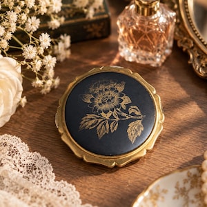 Pode incluir: Um espelho compacto redondo com tampa preta mate e uma ilustração floral em tom dourado. O espelho tem uma moldura metálica de cor dourada com bordas recortadas. O design mostra uma flor central com folhas e caules detalhados.