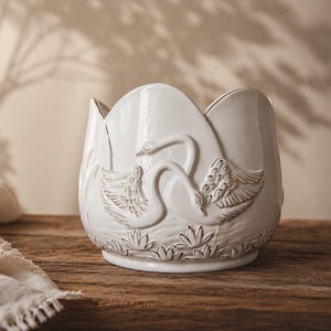 Può includere: Un vaso in ceramica bianca con bordo smerlato e un design in rilievo raffigurante due cigni che nuotano tra le ninfee. La superficie ha una finitura lucida con sottili dettagli anticati che mettono in risalto i motivi scolpiti.