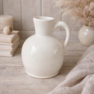 Op de afbeelding: Een witte keramische kan met een afgeronde body en een gebogen handvat. De kan staat op een lichtgekleurd houten oppervlak, met een zachte, neutraal gekleurde doek in de buurt. Decoratieve elementen zijn een kaars en gedroogde planten.